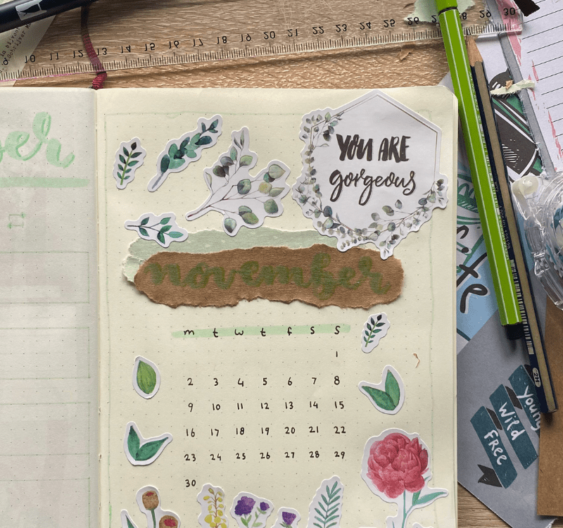 Um bullet journal com vários adesivos de plantas e rosas, um adesivo com a escrita: "you are gourgeous" e o calendário do mës de novembro. 