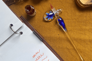 Uma caneta tinteiro de vidro com detalhes em azul. Ao lado da caneta, há um pote pequeno de tinta aberto e um bloco de anotações com folhas brancas e alguns rabiscos sem sentido.