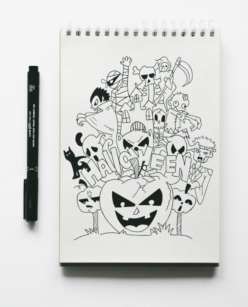 Caneta nanquim preta com detalhes brancos ao lado de um caderno com folhas brancas e um desenho com diversos elementos de halloween.