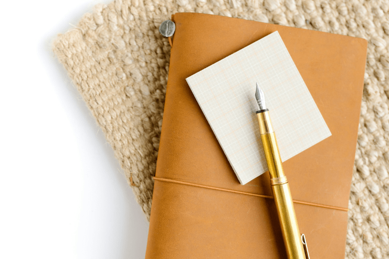 Uma caneta tinteiro dourada em cima de um bloco de papel branco. Ambos estão em cima de um caderno.