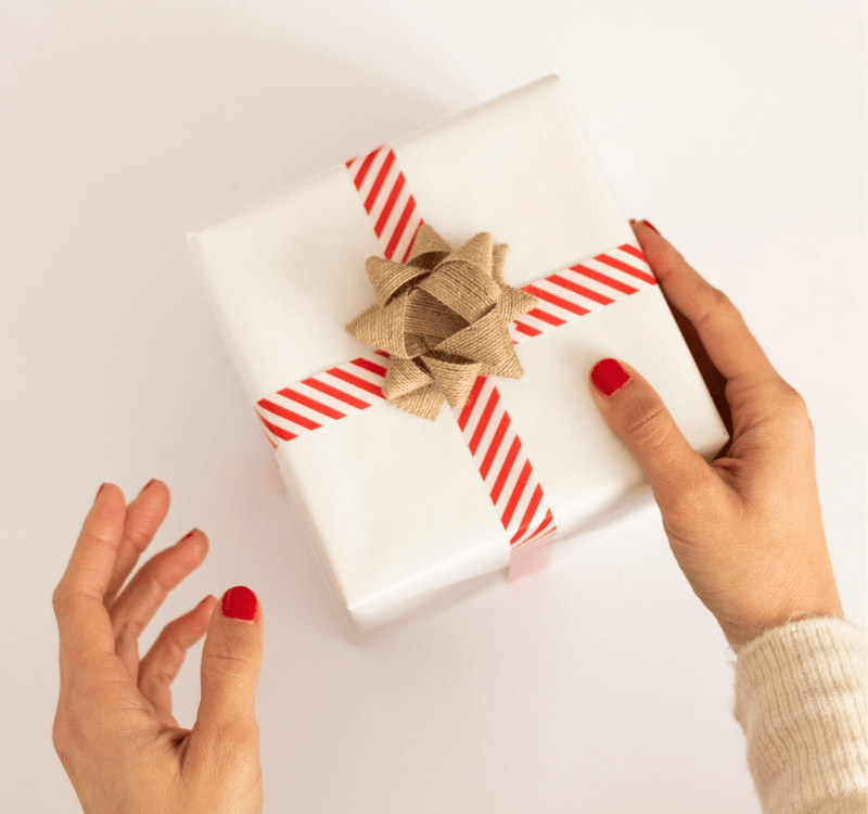 As mãos de uma pessoa branca e unhas pintadas de vermelho, segurando uma caixa de presente embalada em papel branco com fitas tracejadas em vermelho e um detalhe chamativo feito de papel kraft ao centro.