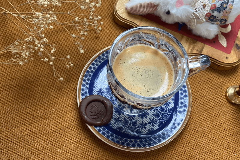 Uma xícara com café no pires e uma moeda de chocolate com detalhes de planta feita com um sinete.