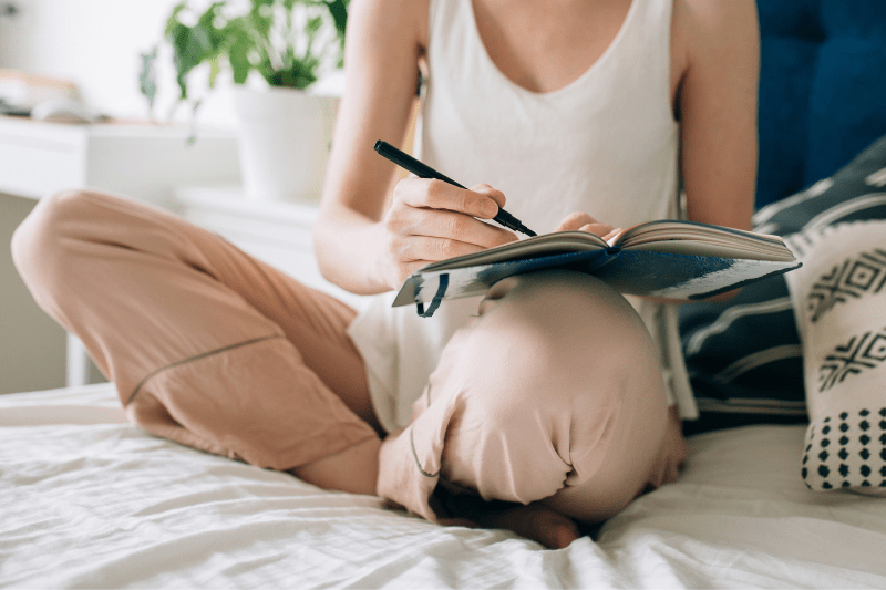 Uma mulher branca sentada em uma cama com um caderno no colo e uma caneta na mão direita.