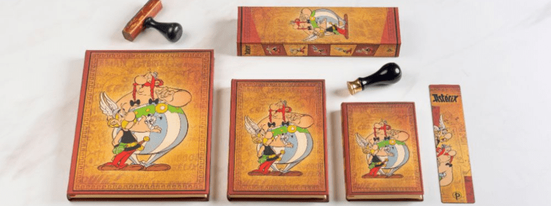 Existem diferentes tamanhos de cadernos para diferentes necessidades. Na foto, temos exemplos da coleção de Asterix & Obelix da Paperblanks com cadernos de diferentes tamanhos, um marcador de página e alguns carimbos espalhados para decorar a foto. Fonte: Paperblanks.